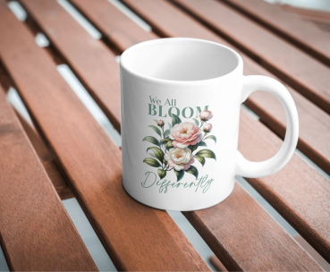 Weiße Keramiktasse mit botanischem Blumenmotiv und dem inspirierenden Zitat „We All Bloom Differently“, fotografiert auf einem Holztisch.